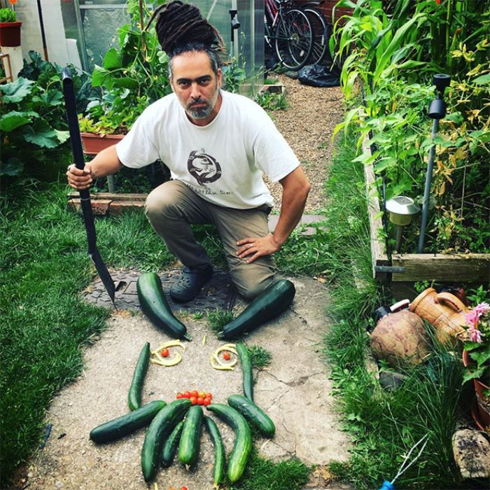 Man with dreadlocks posing as a vegan hunter next to cucumbers arranged like an animal face in a garden setting.