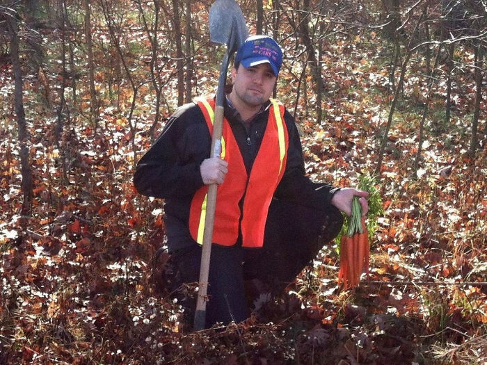 Man dressed in hunting gear holding a shovel and freshly picked carrots in a forest setting for vegan hunters.