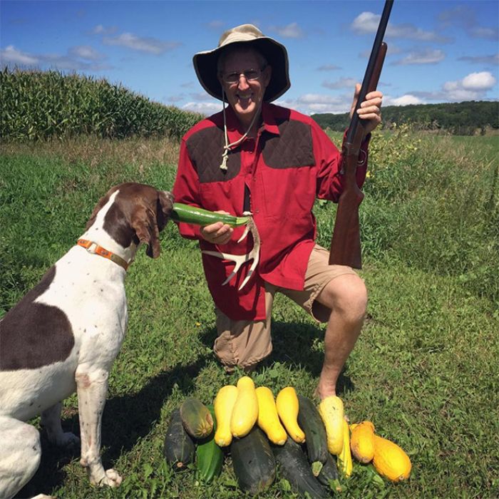 Man dressed as a hunter holding a gun and vegetables with a dog in a field, funny vegan hunters scene.