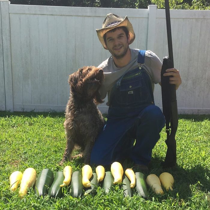 Man in overalls kneeling with dog and holding a toy gun beside a row of zucchini in a funny vegan hunters photo.