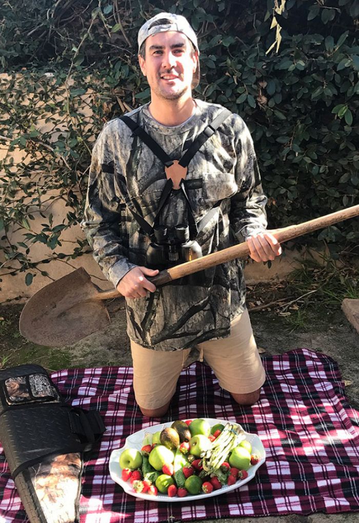 Man dressed for hunting holds shovel over picnic blanket with fresh fruit and vegetables in a humorous vegan hunter scene