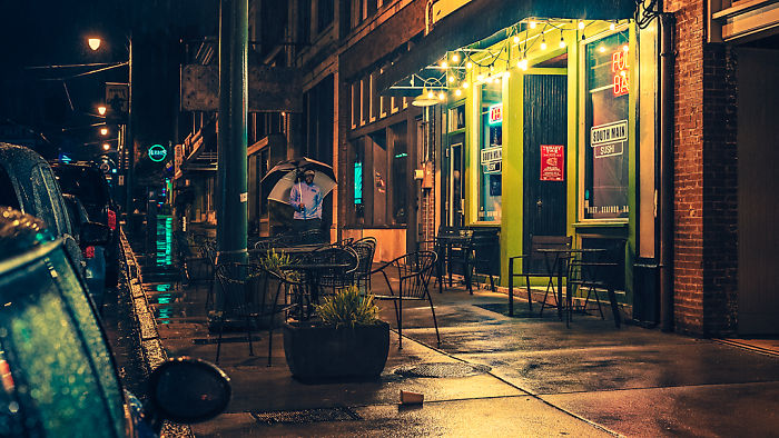 Nighttime street scene in a dangerous city with wet sidewalks, outdoor seating, and a person holding an umbrella near storefronts.