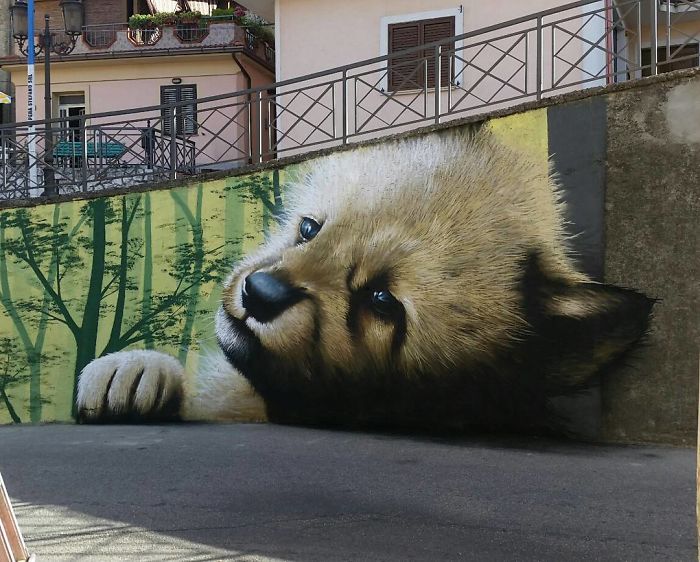 3D street art of a giant panda cub interacting with its surroundings, painted on an urban wall near buildings.