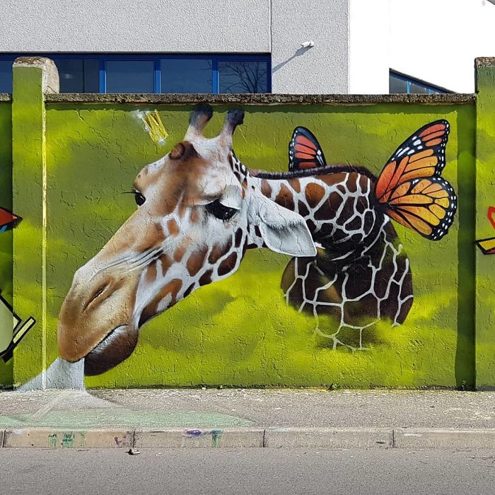 3D street art of a giraffe with butterfly wings painted on an urban wall, interacting with its surroundings.