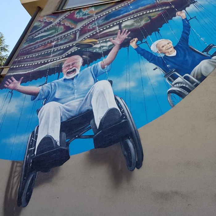 3D street art mural showing elderly people joyfully swinging in wheelchairs, interacting with the building's corner and sky backdrop.