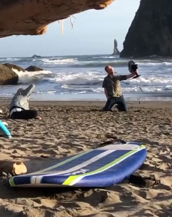 Man taking a picture of another posing on the beach with a fan 