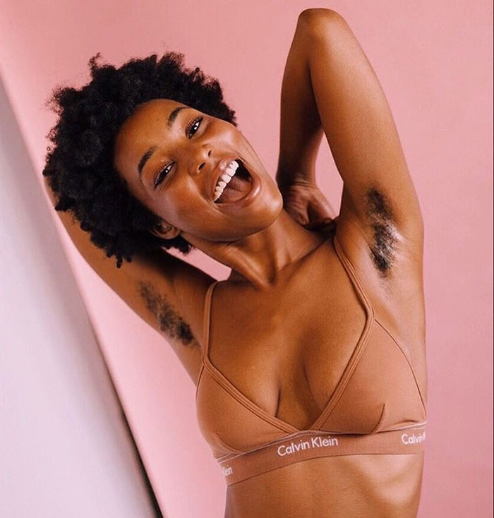 Young woman smiling with natural armpit hair, embracing body positivity and JanuHairy movement against a pink background.