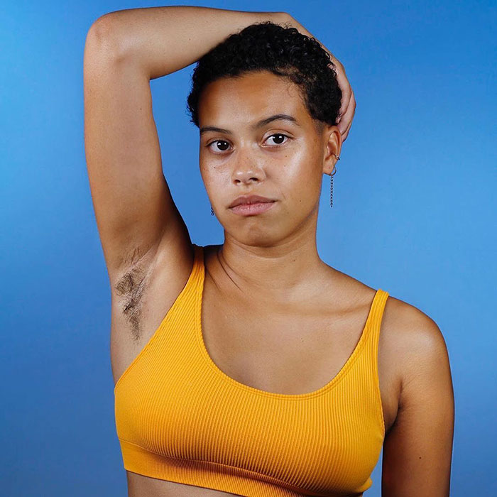 Woman wearing orange top showing natural armpit hair, promoting body positivity and JanuHairy movement against blue background.