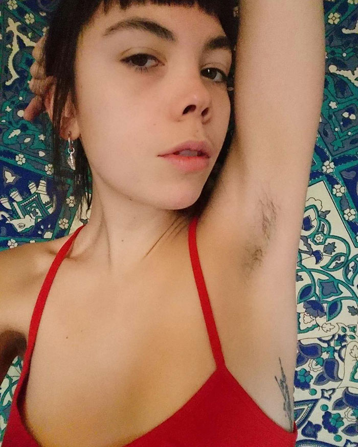 Young woman in a red top showing natural armpit hair against a patterned blue and green background for JanuHairy trend.