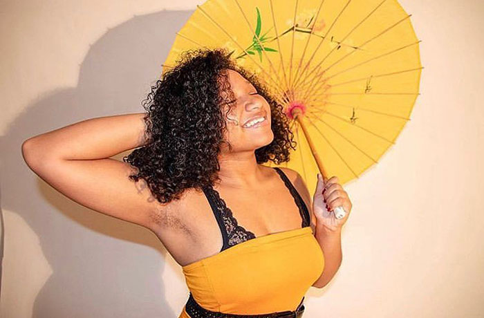 Woman with natural armpit hair smiling and holding a yellow umbrella, embracing body positivity for JanuHairy campaign.