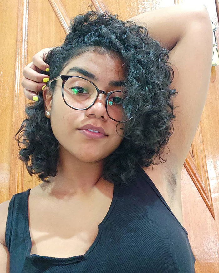 Young woman with curly hair and glasses showing natural armpit hair, embracing JanuHairy body positivity trend.
