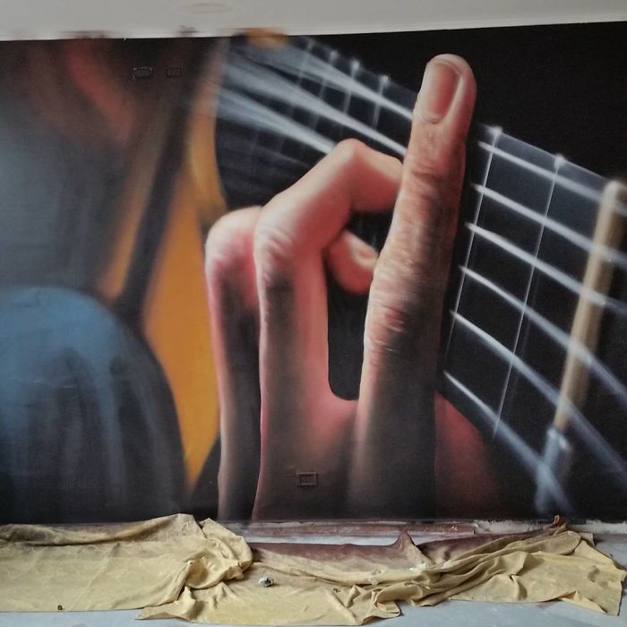 3D street art mural showing a hand playing guitar strings, blending realistically with its surroundings on a wall.