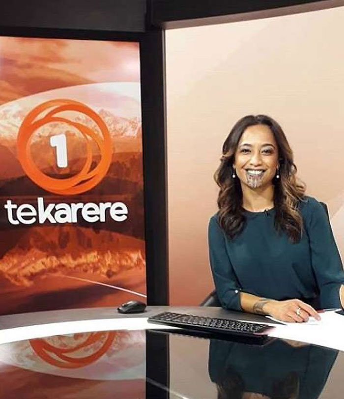 News presenter with a face tattoo smiling at a desk with the Te Karere logo in the background. News presenter with a face tattoo smiling at a desk with the Te Karere logo in the background.