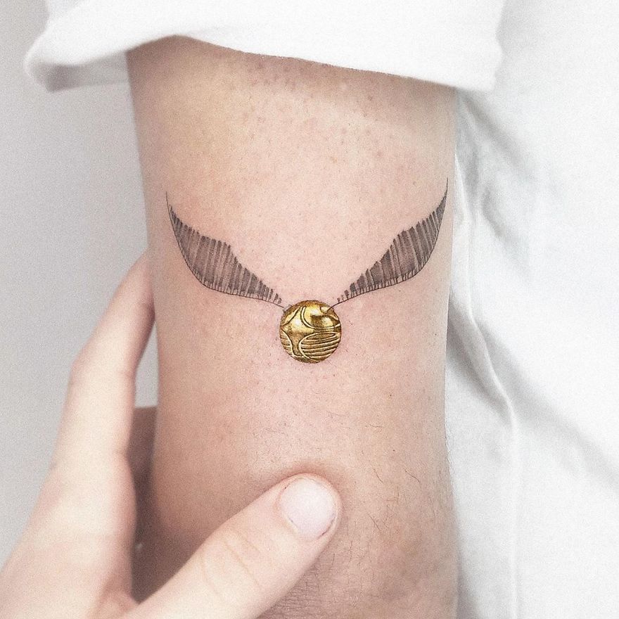 Colorful micro tattoo of a winged golden ball on an arm, resembling a mini pop culture painting by Eden Kozokaro.
