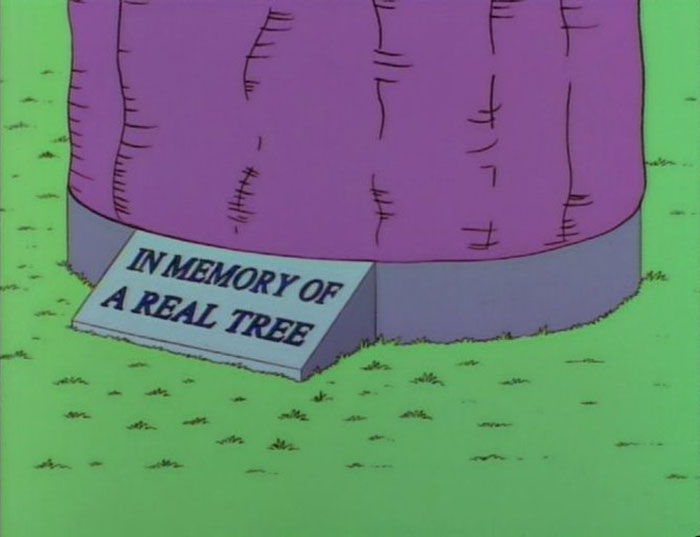 Sign from The Simpsons reading in memory of a real tree placed by a purple cartoon tree stump in grass.