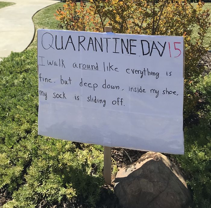 Sign with a dad joke about quarantine day 15, making neighbors laugh. Written in marker, set beside green shrubs.