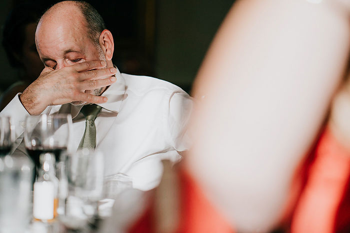 Man in a white shirt wiping tears during unstaged father-daughter moments at wedding reception with blurred guests nearby