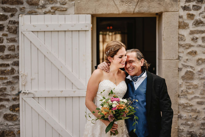 Bride and father sharing a joyful unstaged moment at a wedding, standing outside a rustic stone building.