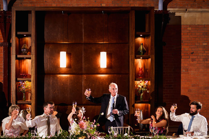 Father giving a heartfelt unstaged wedding toast with daughter and guests raising glasses in celebration.