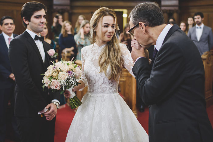 Father gently kissing daughter’s hand during unstaged wedding moment while groom watches in a church setting.