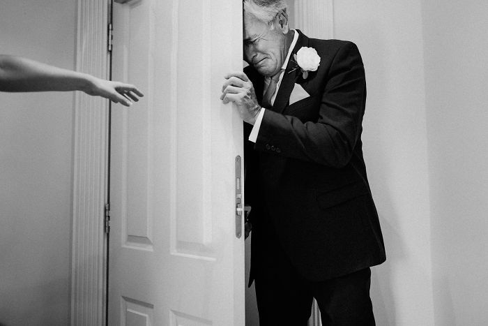 Emotional unstaged father-daughter moment at wedding with father wiping tears behind a door in black and white.