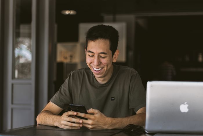 Young man smiling while checking phone next to laptop, depicting employers rejecting job candidates during interview insights.