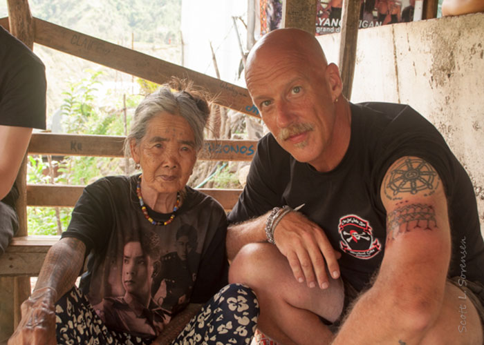 An elderly woman with traditional Filipino tattoos sits beside a man with tattoos in a rustic setting. An elderly woman with traditional Filipino tattoos sits beside a man with tattoos in a rustic setting.