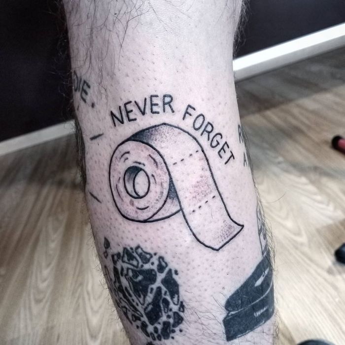 Tattoo of a toilet paper roll with "Never Forget" text, symbolizing a creative Covid-19 design on a person's leg.