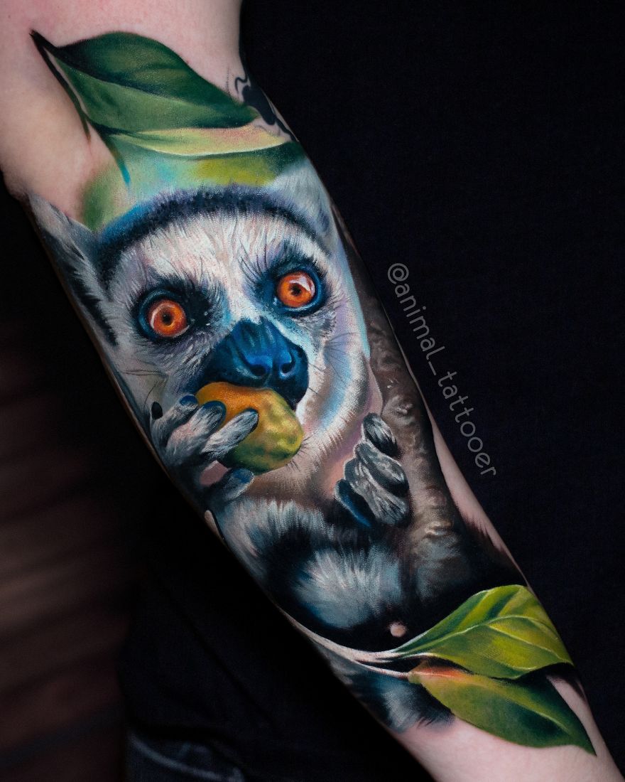 Colorful animal tattoo of a lemur with vibrant orange eyes on the forearm, surrounded by leaves.
