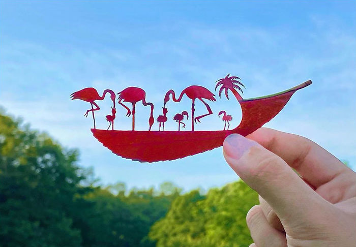 Hand holding a detailed red leaf vignette featuring flamingos and a palm tree against a blue sky and greenery.