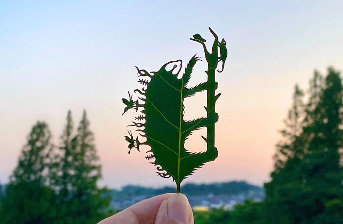 Hand holding a leaf art vignette with detailed cutouts by Japanese artist against a soft sky and tree background.