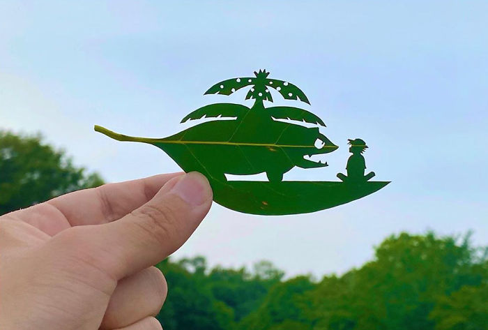 Hand holding a leaf with detailed cutout vignette of a creature and person, showcasing Japanese artist leaf art.