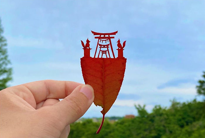 Hand holding a red leaf with a cute vignette of a Japanese shrine and fox statues cut from the leaf.
