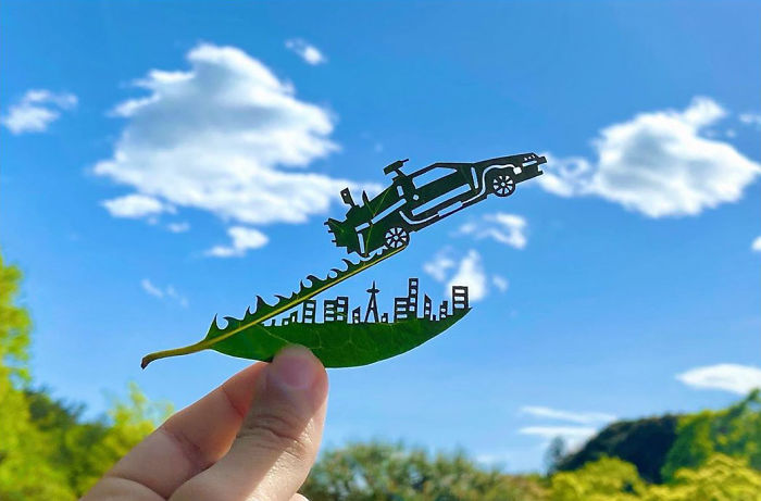Hand holding a leaf cut into a vignette with a cityscape and car against a blue sky by Japanese artist.