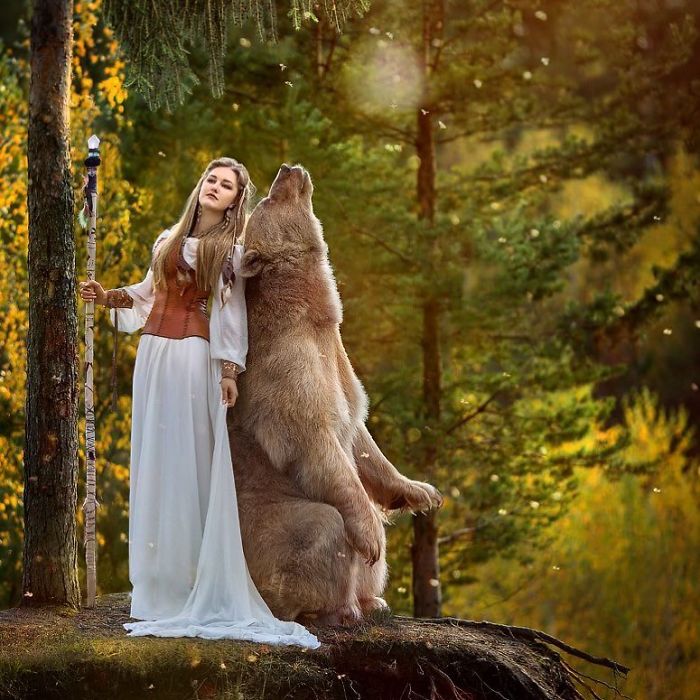 Young woman in a white dress posing in a forest with a Russian brown bear standing upright beside her.