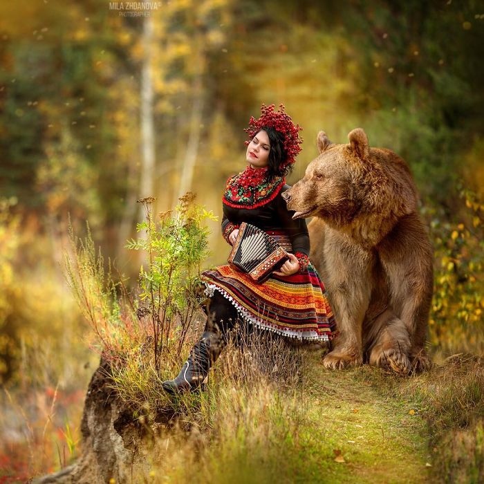 Woman in traditional clothing playing an accordion beside a Russian brown bear in a vibrant autumn forest setting.