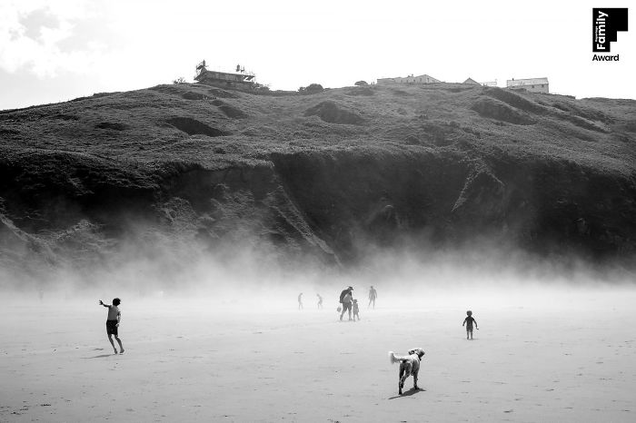 Black and white award-winning family photo capturing a misty beach moment with children and a dog near a hillside.