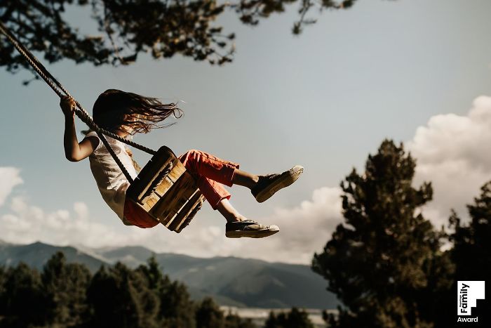 Child swinging outdoors with hair flying, capturing a family moment in an award-winning photo against a scenic mountain backdrop.