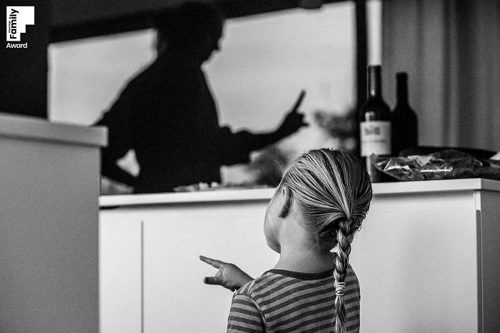 Child with braided hair pointing while an adult is silhouetted in the background, capturing a candid family moment photograph.