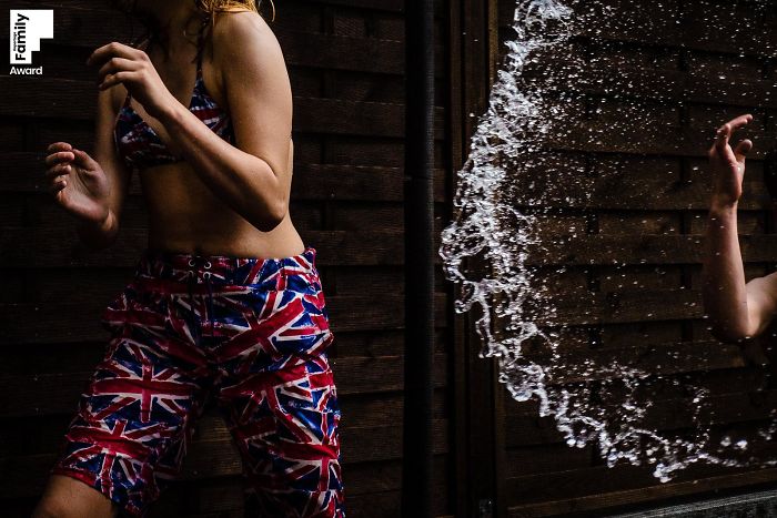 Child playing with water splash wearing Union Jack shorts, capturing the art of a family moment in an award-winning photo.