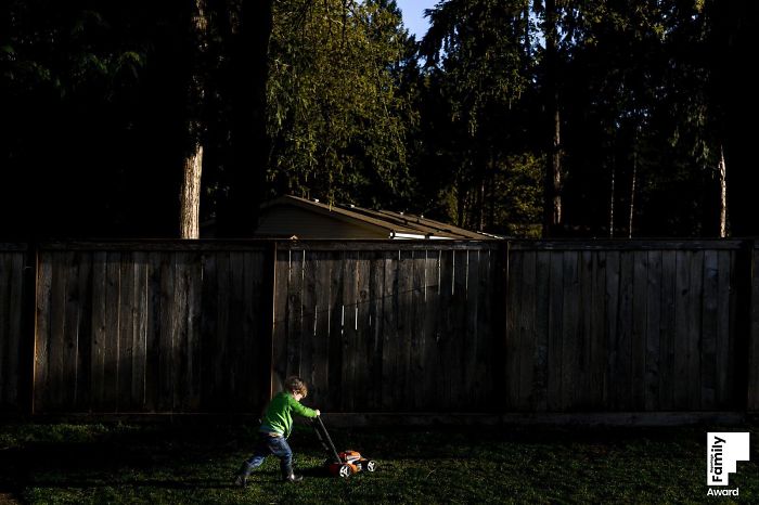 Child in a green shirt pushing a toy lawn mower in a backyard, capturing a family moment in natural light.