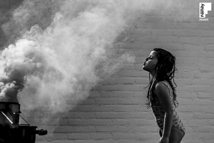 Black and white award-winning family photo of a young girl interacting with steam against a brick wall background.
