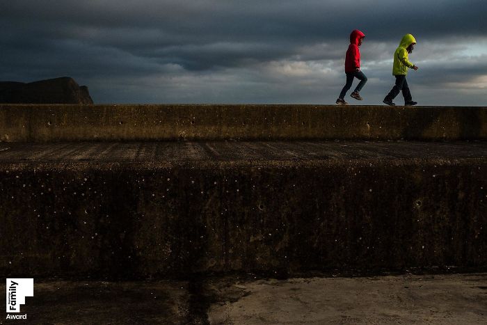 Two children wearing red and yellow jackets walk on a concrete wall, capturing a family moment in moody natural light.