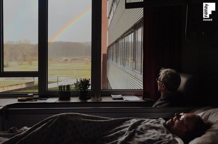 Man sitting by window looking at rainbow while another person rests in bed, capturing a family moment award-winning photo.