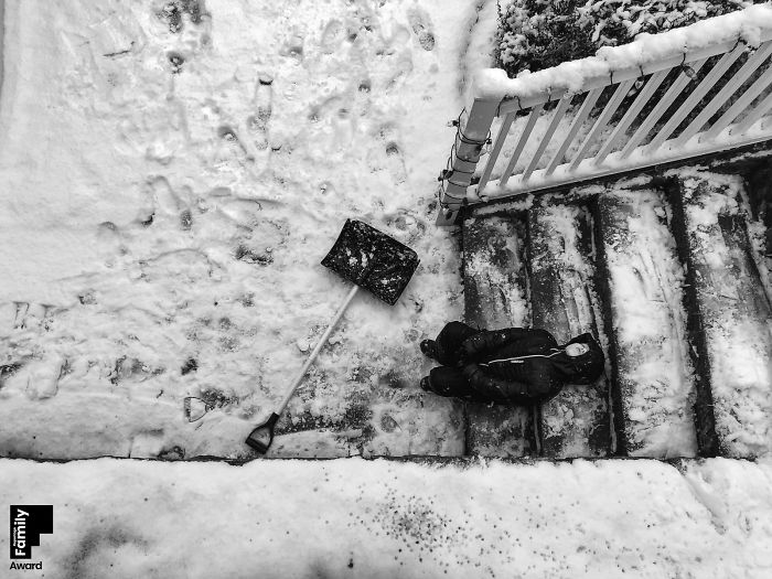 Black and white photo capturing a family moment with a child resting on snowy outdoor stairs next to a snow shovel.