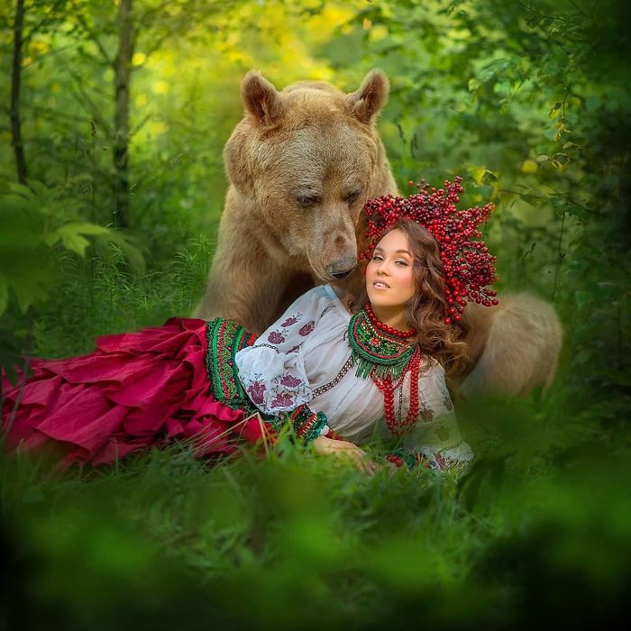 Russian brown bear and woman in traditional attire posing together in a lush green forest for a modeling shoot.