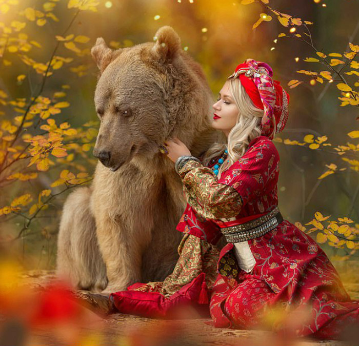 Woman in traditional Russian attire sitting beside a Russian brown bear in an autumn forest setting.