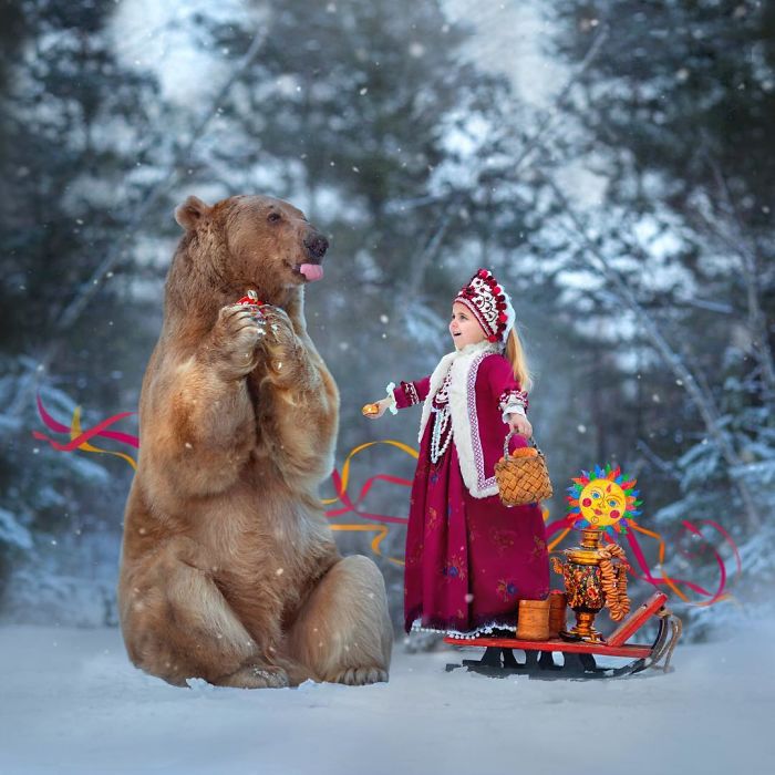 Russian brown bear sitting in snow with a young girl in traditional dress during a winter outdoor scene.
