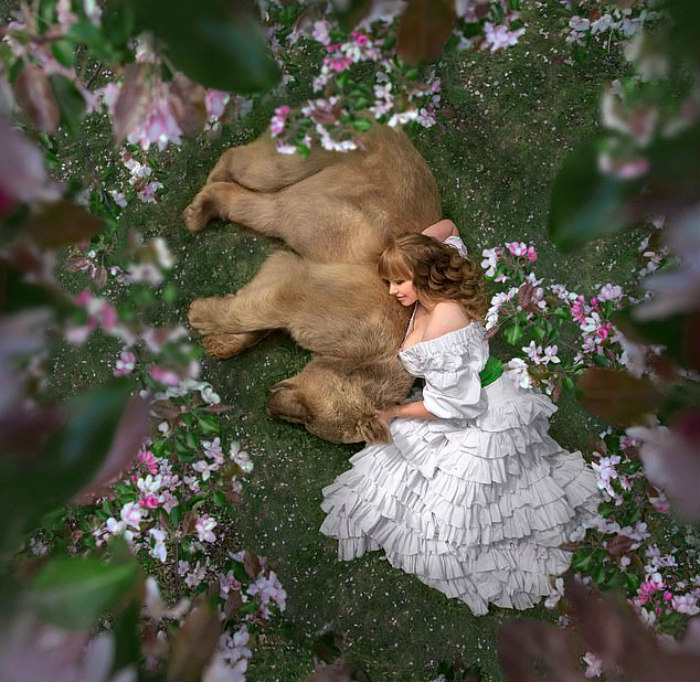 Young woman in a white dress lying beside a Russian brown bear cub surrounded by pink flowers outdoors.