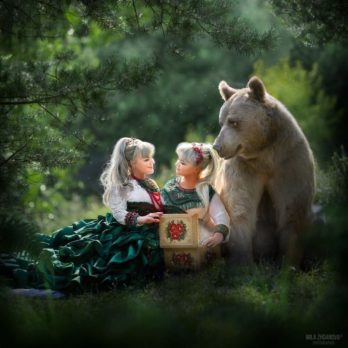 Russian brown bear sitting calmly beside two girls in traditional dresses in a lush green forest setting.